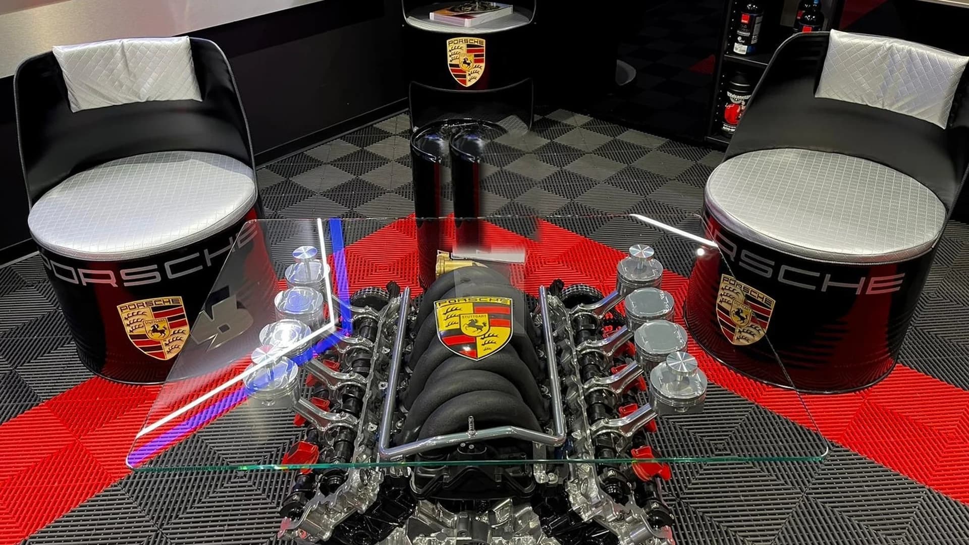 Garage aménagé par ADS Concept Store : table basse sur bloc moteur V8 à plateau de verre, fauteuils sur fûts laqués et dalle de sol en damier rouge et noir